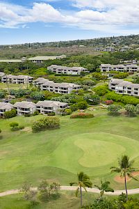 Aerial view of a golf course with a green and fairways, surrounded by low-rise apartment buildings and lush greenery, under a blue sky.