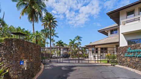 A gated entrance to a tropical resort with palm trees, stone walls, and a two-story white building labeled &ldquo;Hawaiian &hellip;&rdquo; at the gate; sunny and blue skies.
