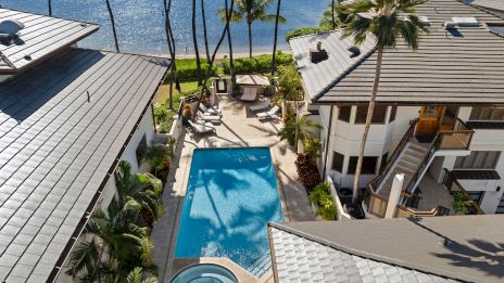 Luxurious beachfront resort with a long rectangular pool, hot tub, palm trees, and ocean views between two gray-roofed buildings.