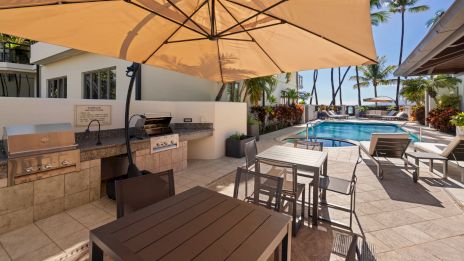 A resort-style poolside patio with outdoor seating, a built-in grill/BBQ area, sun loungers, palm trees, and a large beige umbrella shading tables.