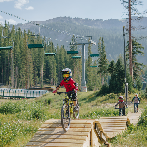 The image shows children riding bicycles on a wooden pathway in a scenic outdoor area with chairlifts, trees, and mountains in the background.