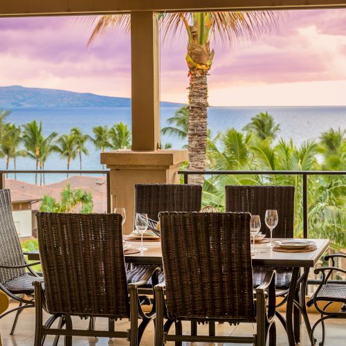 A dining setup on a balcony overlooking a tropical landscape with palm trees and ocean views during sunset.