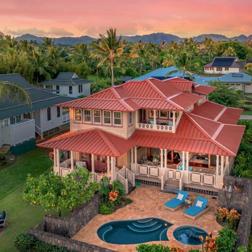 A beautiful, two-story house with a red roof, a pool, lounge area, and tropical scenery with other houses and mountains in the background.