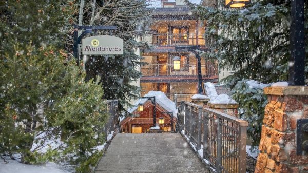 A snowy walkway leads to a cozy, warmly lit lodge surrounded by trees and a sign reading "Antantonares" hangs on the left.