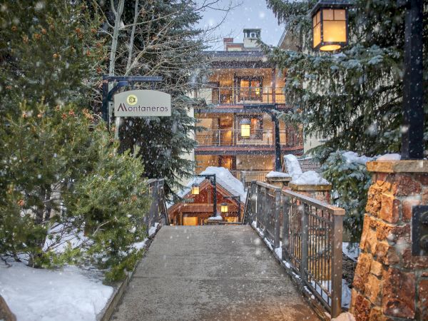 A snowy walkway leads to a cozy, warmly lit lodge surrounded by trees and a sign reading 