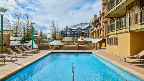 The image shows a serene pool area surrounded by lounge chairs, with mountains and buildings in the background under a clear sky.