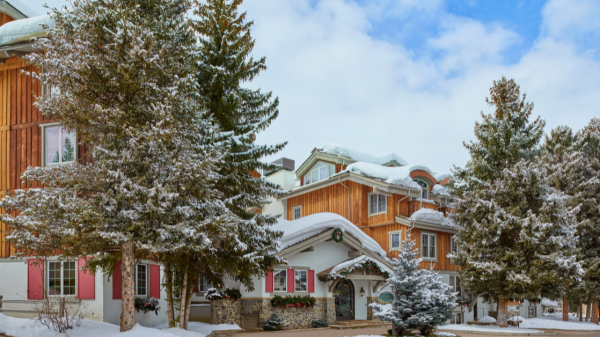 The image depicts a snow-covered building surrounded by evergreen trees under a cloudy blue sky. It's a wintery scene.