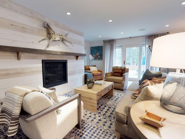 A cozy, modern living room with a fireplace, various seating, a coffee table, and a lamp. Large windows bring in natural light, enhancing the decor.