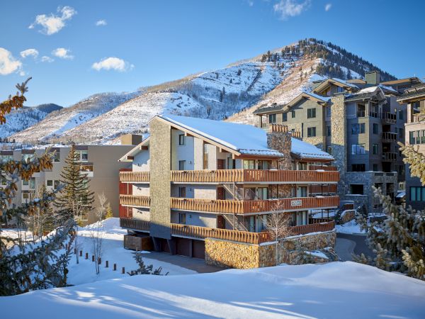 Snow-covered buildings with balconies, surrounded by evergreen trees, set against a mountainous backdrop under a blue sky with scattered clouds.