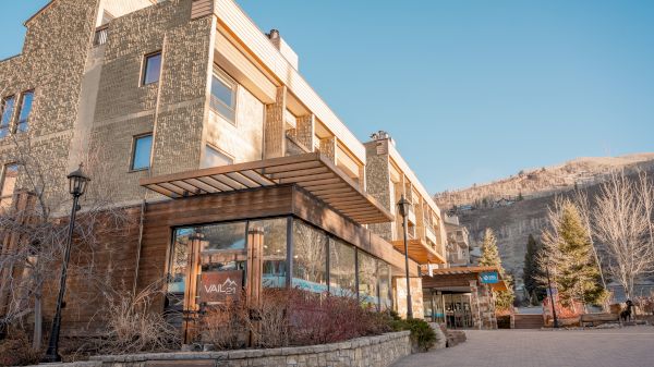 The image shows a modern stone building with glass windows, surrounded by trees and a clear blue sky, creating a serene atmosphere.
