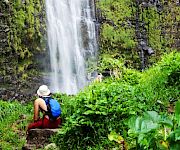 A person with a blue backpack and white hat sits on a rock, admiring a waterfall surrounded by lush green vegetation.