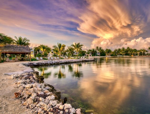 A serene lakeside scene with a sandy shore, lush palm trees, and dramatic clouds reflecting in the water at sunset.