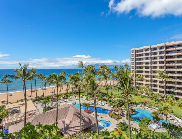 A beachfront hotel with a large pool area surrounded by palm trees, overlooking the ocean under a sunny blue sky.