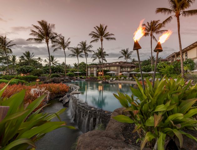 A tropical resort scene with palm trees, a pool with a waterfall, lush greenery, and tiki torches lit at dusk.