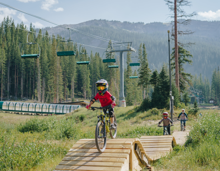 Three people are mountain biking on a wooden trail in a scenic location with lift chairs and trees in the background, under a clear sky.