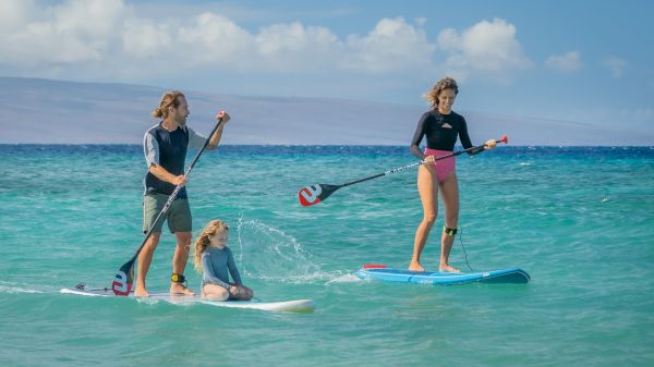 A family enjoys stand-up paddleboarding in clear blue water, with a sunny sky and gentle waves in the background.