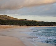 A scenic beach with calm waves, a few people relaxing on the sand, lush greenery, and hills under a partly cloudy sky.