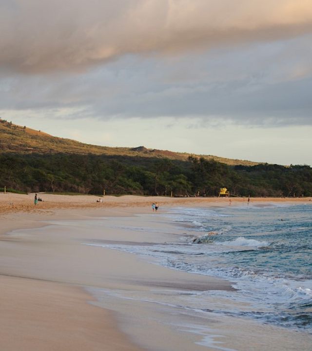 A scenic beach with calm waves, a few people relaxing on the sand, lush greenery, and hills under a partly cloudy sky.