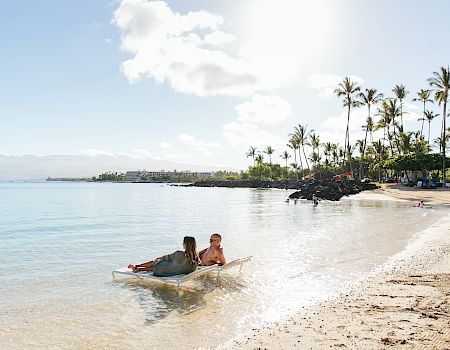 Two people are lounging on beach chairs partially submerged in clear, calm water, with a sandy beach and palm trees in the background.