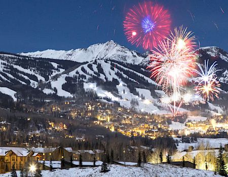 A nighttime winter scene with lit buildings in the foreground, snowy mountains in the background, and colorful fireworks in the sky, ending the sentence.