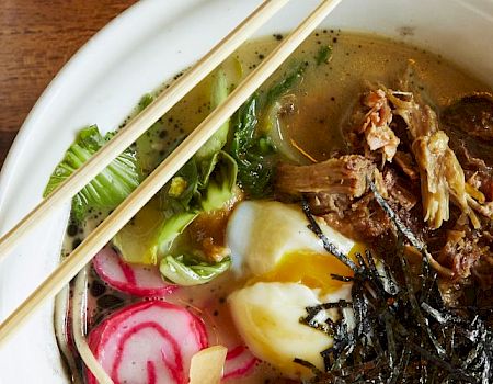 Two bowls of Asian noodle dishes, one with lime wedges and crispy toppings, the other with shredded meat, radishes, and seaweed.