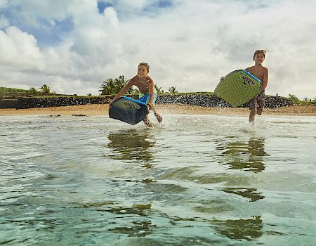 Two kids are running through shallow water, carrying boogie boards on a sunny beach day. They appear excited and playful.