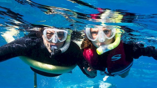 Two people are snorkeling underwater, wearing masks and fins, smiling and enjoying their time in the clear blue water.
