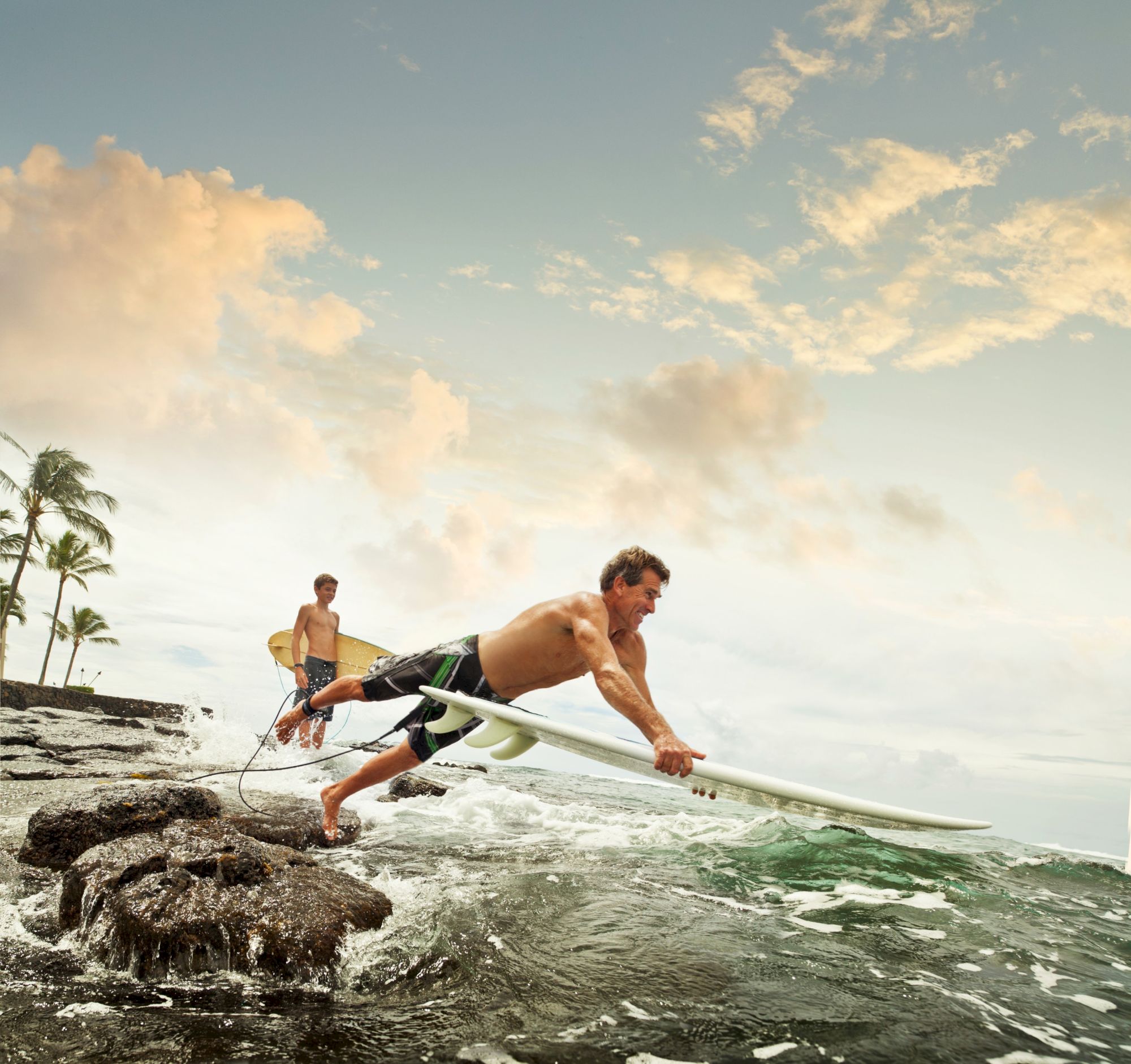Two surfers catching a wave along a rocky shoreline at sunset, one on the board leaning forward while another follows behind, palm trees in the background.