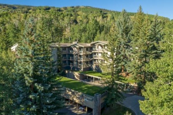 The image shows a multi-story building surrounded by lush trees and mountainous terrain under a clear blue sky.