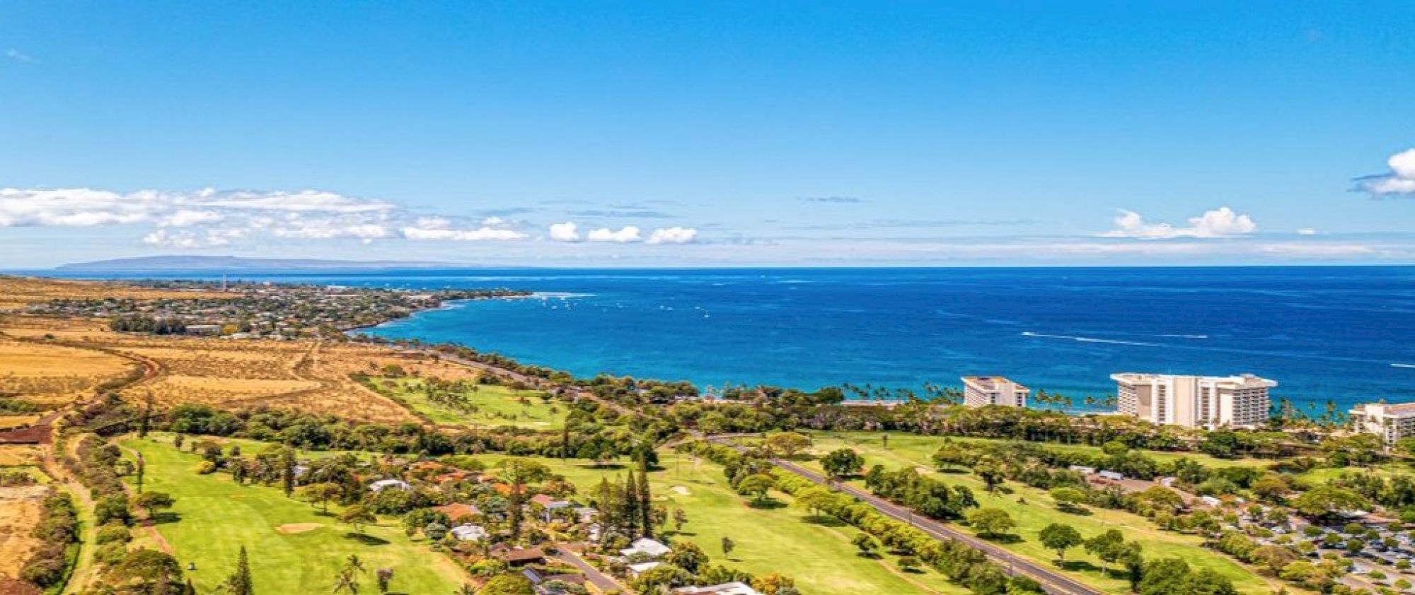 The image shows a scenic coastal view with lush greenery, hills, and a clear blue ocean under a bright sky.