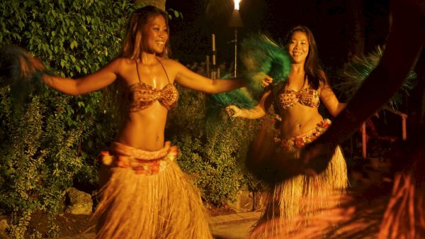 The image shows two women dancing in traditional attire with grass skirts and vibrant accessories, illuminated in a warm, inviting glow.