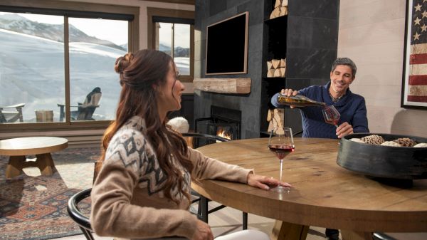 A man pours wine for a woman in a cozy room with a fireplace and snowy views, creating a warm, inviting atmosphere.