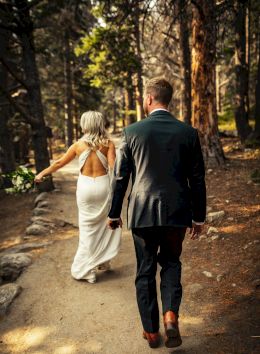 A couple walks hand-in-hand along a forest path, the woman in a white dress and the man in a suit, surrounded by nature.
