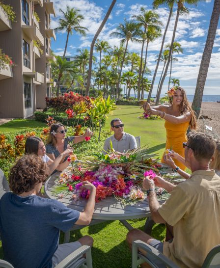 A group of people enjoys a sunny day outdoors, crafting with colorful flowers near the beach and tropical scenery.