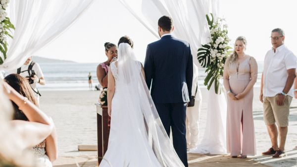 A couple is exchanging vows at a beach wedding, with a decorated arch, surrounded by guests enjoying the beautiful setting.