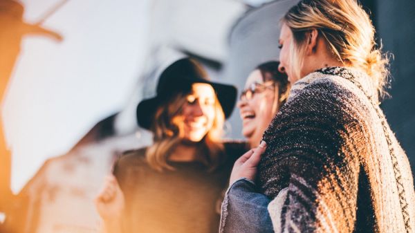 The image shows three women laughing and enjoying each other's company outdoors, wrapped in cozy attire.