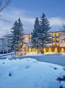The image shows a cozy hotel or lodge surrounded by snow-covered trees, illuminated at dusk. A serene winter scene unfolds.