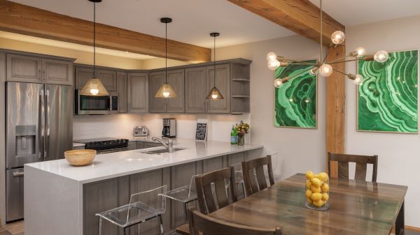 The image shows a modern kitchen with wooden beams, sleek cabinets, and a dining area featuring a table with lemons.