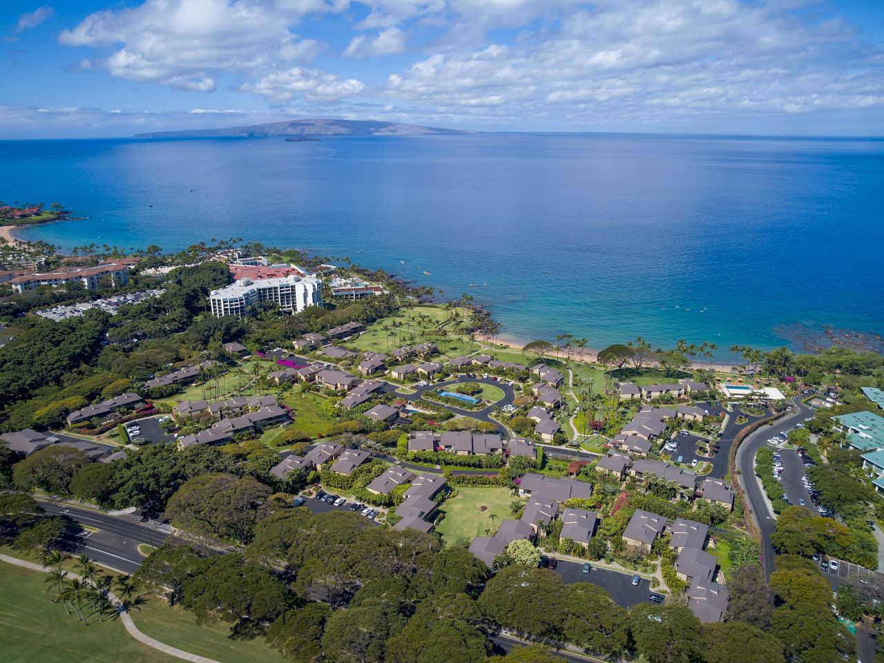 The image shows a coastal view featuring lush greenery, residential areas, and a serene ocean landscape under a blue sky.