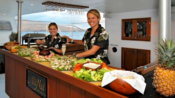 Two women in floral shirts prepare a colorful array of food on a boat, with a scenic view visible in the background.