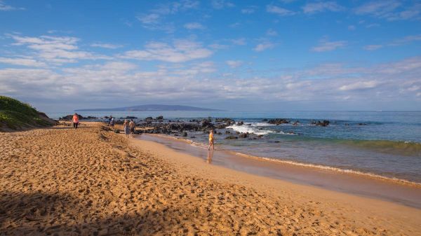 The image shows a sandy beach with gentle waves, rocky formations, and a clear blue sky, creating a peaceful coastal scene.