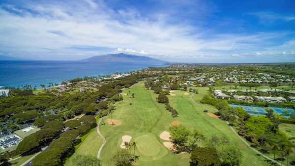 The image shows a scenic view of a golf course by the ocean, with lush greenery and distant mountains under a blue sky.
