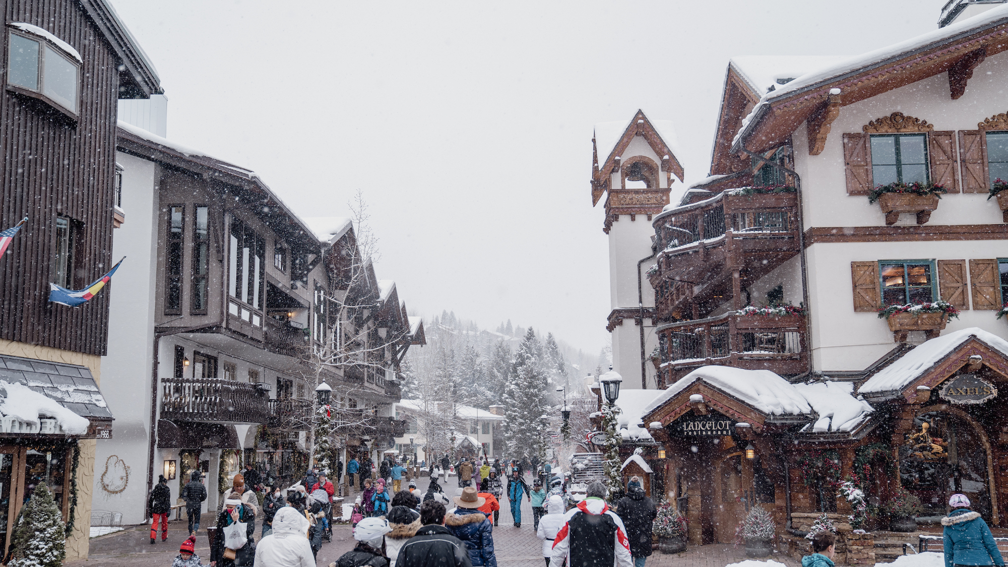 A snowy street scene with people walking among charming alpine-style buildings and shops, creating a festive winter atmosphere.