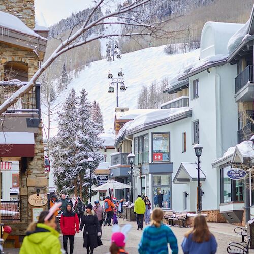 The image shows a snowy street with shops and people walking, surrounded by winter scenery and ski slopes in the background.