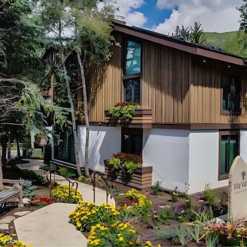 The image shows a modern building surrounded by trees, colorful flowers, and a pathway leading to it. It has a welcoming appearance.