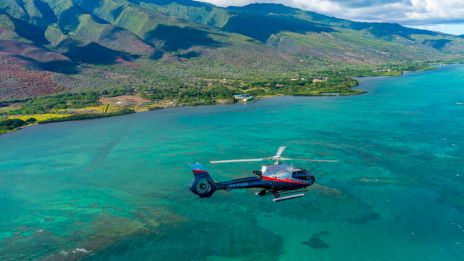 A helicopter flies over a vibrant turquoise ocean, with lush green mountains and coastline in the background. A scenic view unfolds.