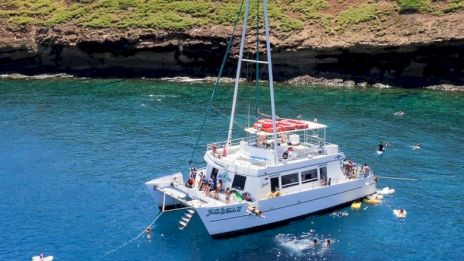 The image features a boat anchored in clear blue water near a rocky shoreline, with a few people nearby enjoying the scene.