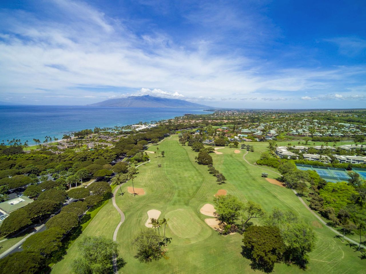 The image shows a scenic aerial view of a golf course near the coast, with lush greenery and a distant mountain backdrop.