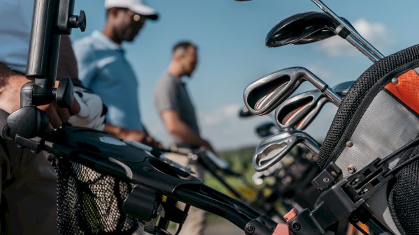 The image shows a group of golfers near their golf bags, with clubs visible against a clear blue sky.
