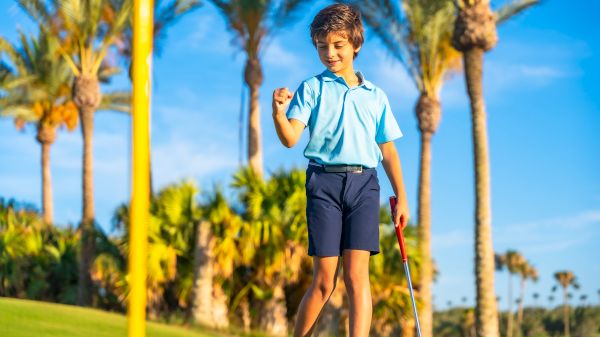 A young boy in a blue golf shirt and shorts walks on a green, smiling brightly amid palm trees and a sunny sky.
